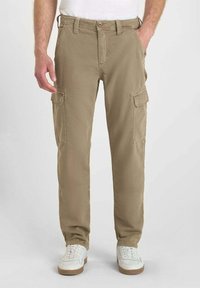 Man som bär beiga cargobyxor med fram- och sidofickor samt vita sneakers, stående mot en enfärgad bakgrund.