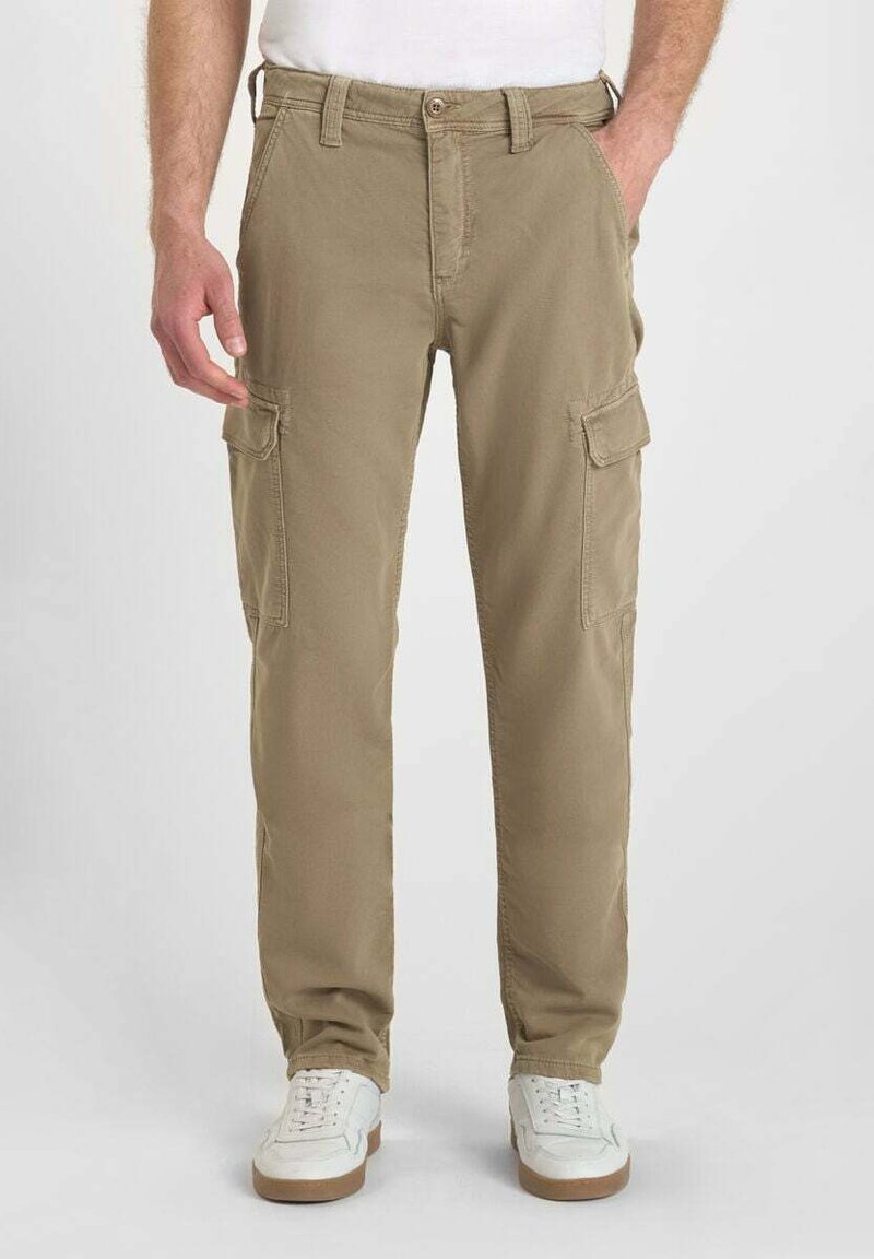 Man som bär beiga cargobyxor med fram- och sidofickor samt vita sneakers, stående mot en enfärgad bakgrund.