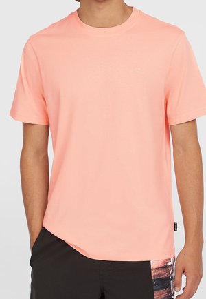 Mężczyzna ubrany w jednokolorowy jasnoróżowy t-shirt z krótkim rękawem i czarne szorty z graficznym wzorem po jednej stronie, stojący z jedną ręką w kieszeni.