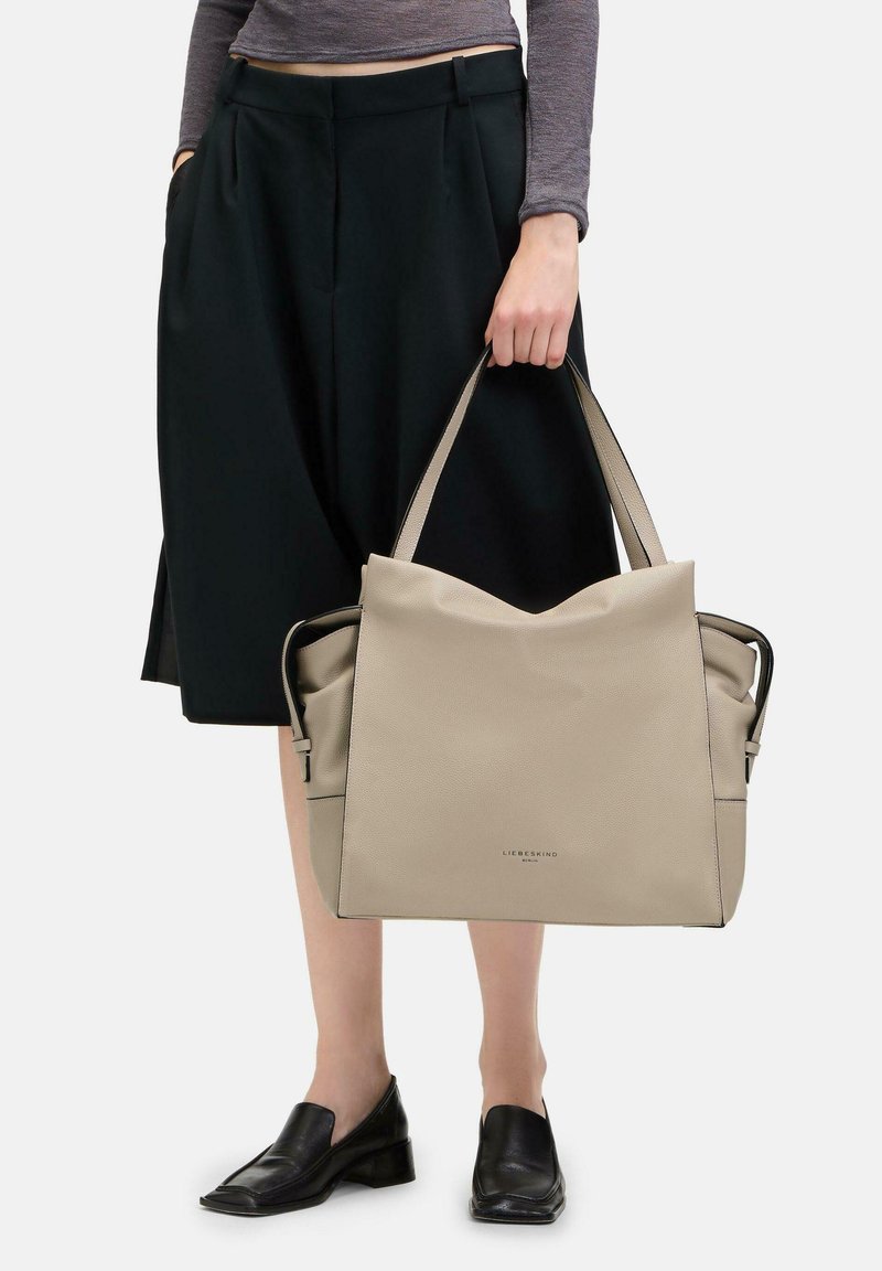 Sac tote en cuir beige avec un design décontracté et double anses, présentant une surface texturée et un logo de marque subtil à l'avant.