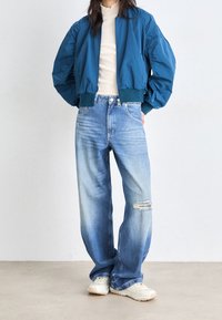 Teal bombarjacka med puffärmar, tillsammans med ljusblå slitna jeans med ett trasigt knä och vita sneakers. Enkel texturerad design.