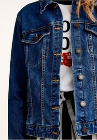 Veste en jean avec boutons en métal portée sur un t-shirt graphique blanc et un pantalon rayé noir et blanc noué à la taille.