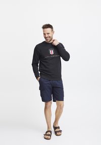 Felpa nera con logo rosso e bianco, abbinata a pantaloni cargo blu navy e sandali marroni. Texture liscia, vestibilità casual e design semplice.