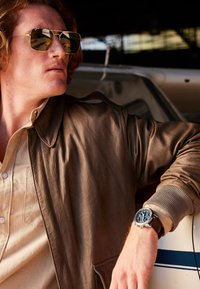 Veste aviateur en cuir marron avec des poignets côtelés, chemise beige et montre-bracelet en argent avec un cadran bleu. Des lunettes de soleil dorées réfléchissantes complètent le look.