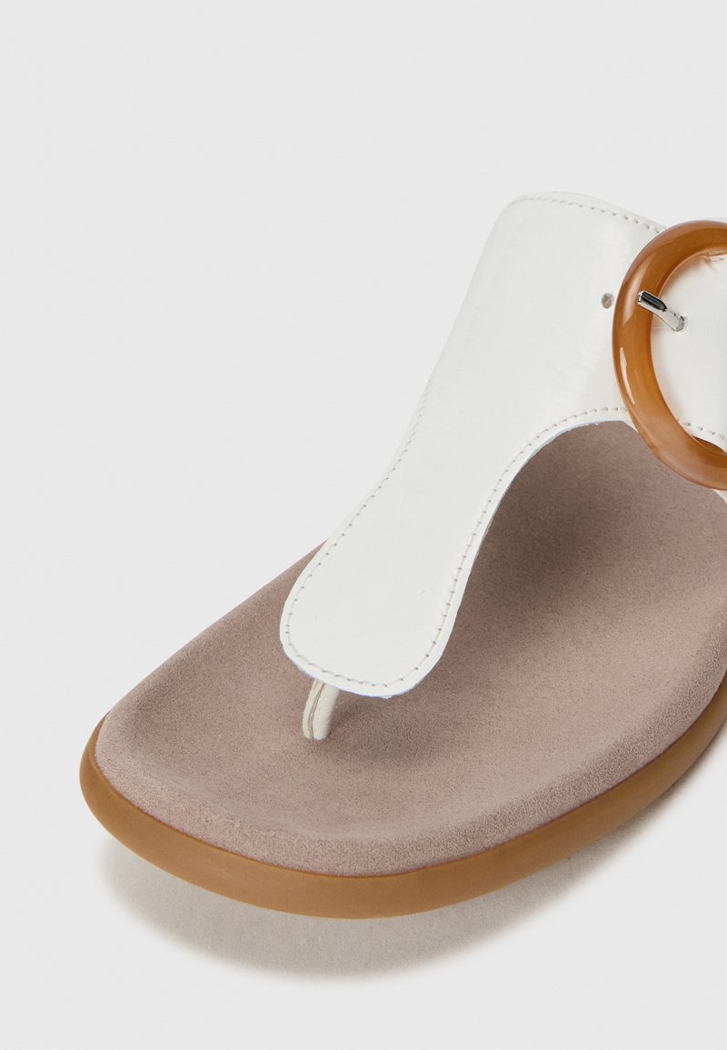 Witte t-bar sandaal met een grote ronde bruine detail, rustend op een zacht beige oppervlak, waardoor het slanke ontwerp wordt benadrukt.