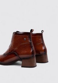 Botas de tobillo de cuero marrón con un acabado brillante, que cuentan con un tacón cuadrado y lengüetas en los laterales para facilitar su uso.