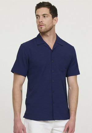 Homme portant une chemise à manches courtes boutonnée bleu marine texturée avec un col et une poche avant, associée à un pantalon blanc.