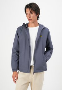 Jeune homme portant une veste à capuche zippée bleue avec des poignets élastiques, un t-shirt blanc et un pantalon beige, se tenant devant un fond blanc uni.