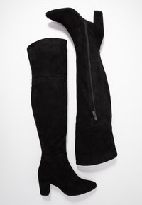 Pari mustia mokkanahkaisista polvipituisista saappaista, joissa on paksut korot ja sivuvetoketjut, aseteltuna tasaisena valkoista taustaa vasten.