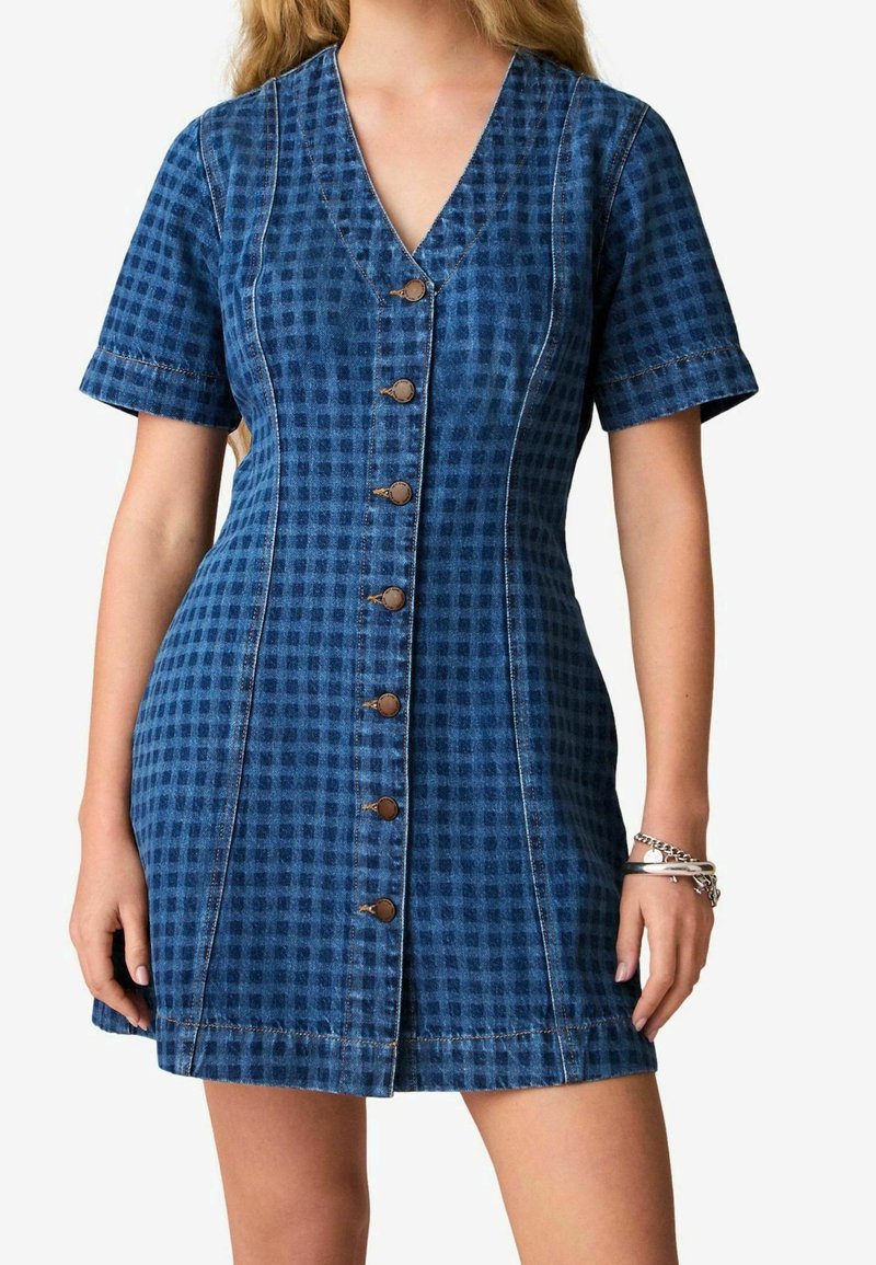 Robe en denim ajustée à manches courtes bleue à carreaux avec boutons sur le devant, col en V et bracelets argentés au poignet droit.