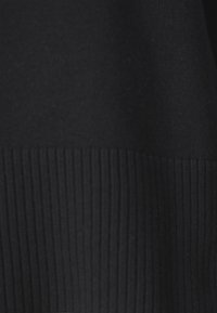Le tissu du pull en maille noir présente une texture lisse avec un motif côtelé en bas. La couleur est uniforme sans autres détails notables.