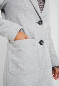 Grey wool coat with a tailored design, two buttons, a textured fabric, and a large pocket on the front. Accented by a knitted cuff.