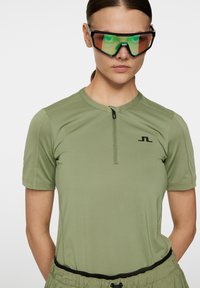 Lyhythihainen, oliivinvihreä urheilupaita, jossa on puolivetoketjukaulus ja logoyksityiskohta. Henkilöllä on päällä mustat ja vihreät urheilulasit. Pintamateriaali on sileä.