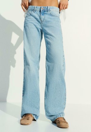 Personne portant un jean large bleu clair avec des boutons latéraux et des mocassins marron, debout devant un fond blanc.