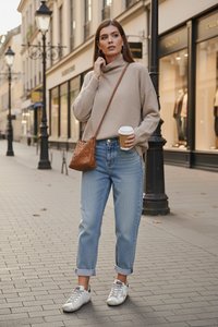 Žena v béžovom svetri a modrých džínsoch drží šálku kávy a hnedú crossbody tašku, stojí na mestskom chodníku blízko obchodov.