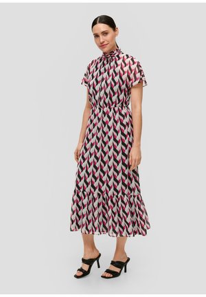 Frau trägt ein gemustertes Midi-Kleid mit rosa, schwarz und weißen geometrischen Formen, kombiniert mit schwarzen Absatzsandalen, stehend vor einem schlichten Hintergrund.