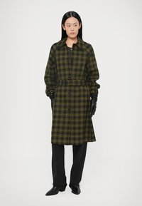 Green and black plaid long shirt dress with a belted waist, button closure, and a collar. Worn with black gloves and black pants.