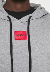 Sudadera gris con un parche rectangular rojo que lleva el texto "HUGO". Cuerdas negras, tela texturizada y un ajuste relajado.