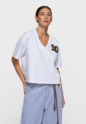 Biały krótki top z dekoltem w serek i brązowym haftowanym detalem "10" po lewej stronie, wykonany z miękkiego bawełnianego materiału. W zestawie z pasiastymi spodniami.