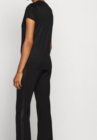 Camiseta negra de manga corta en tejido suave, con un escote redondo. Llevada con pantalones negros que tienen una franja satinada en el lateral.