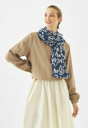 Vrouw draagt een beige jas, crèmekleurige rok en een marineblauwe sjaal met witte abstracte letters, glimlachend en zijwaarts kijkend, handen op de heupen.