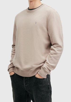 Mand iført beige crewneck sweatshirt med lille brystlogo, hænderne i lommerne, kombineret med sorte jeans, vist fra nakke til hofter.