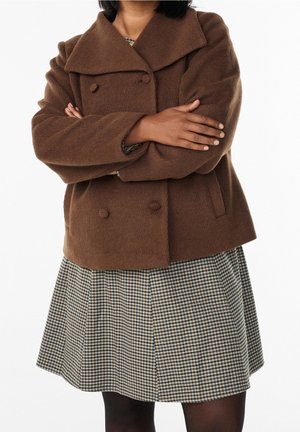 Personne les bras croisés portant un manteau marron à double boutonnage et une jupe à carreaux par-dessus des collants noirs sur fond blanc.