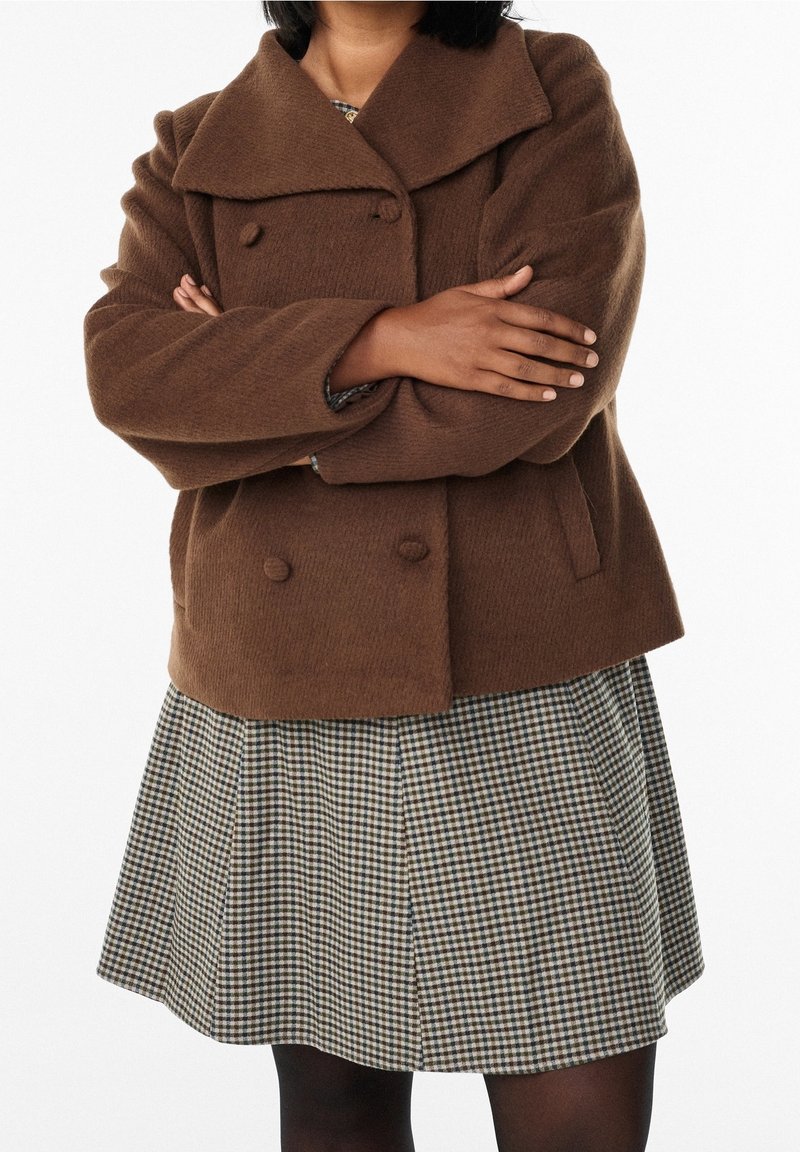 Personne les bras croisés portant un manteau marron à double boutonnage et une jupe à carreaux par-dessus des collants noirs sur fond blanc.