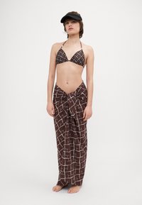Brun og hvid mønstret bikini top med trekantede skåle, parret med en matchende, bundet sarongkjole. Modellen bærer en sort skyggehue.