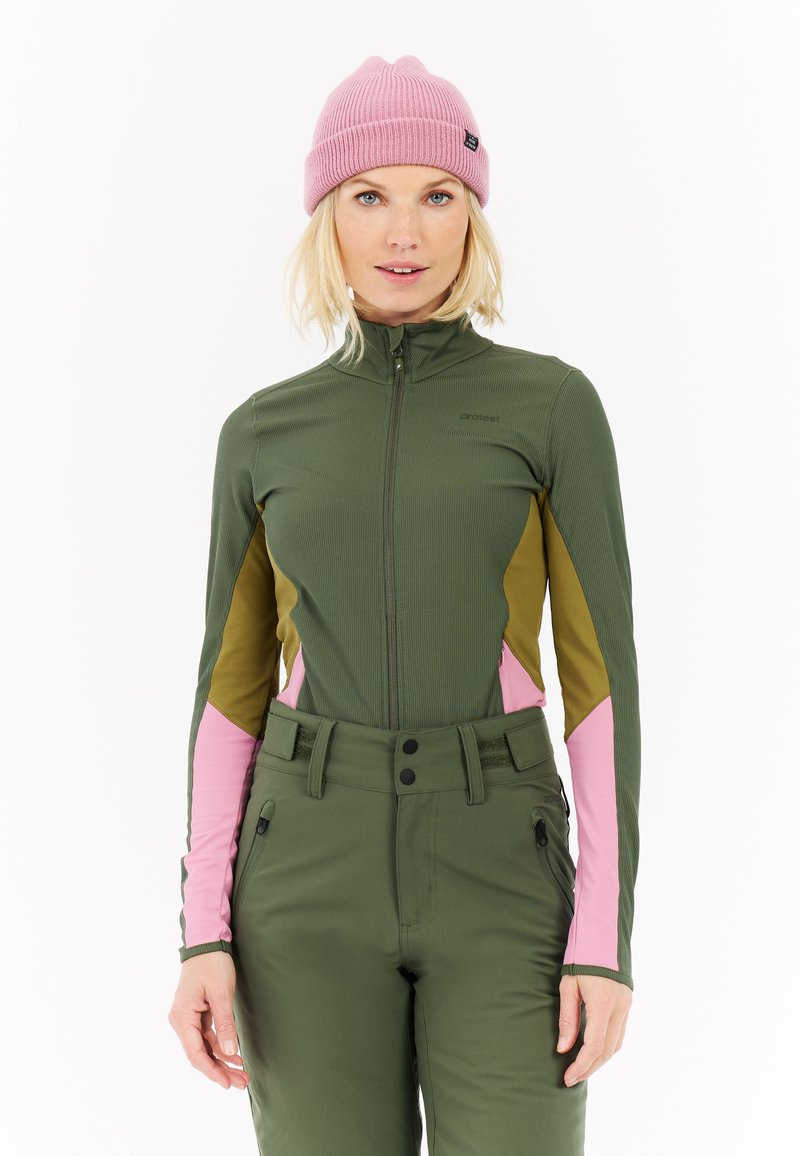 Žena v olivovo zelenom zipsovom saku s ružovými a hnedými panelmi, zodpovedajúcich olivovo zelených nohaviciach a ružovej pletenej čiapke, stojí pred bielym pozadím.