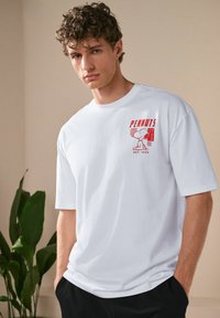 White short-sleeve t-shirt made of cotton, featuring a red graphic print of a character on the left chest. Relaxed fit and crew neck design.