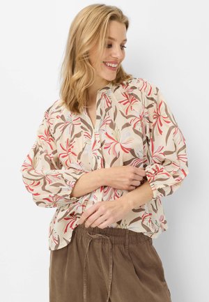 Femme souriante ajustant une blouse crème à motif floral rouge et marron, associée à un pantalon marron ample à cordon, debout sur fond blanc.