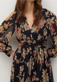 Woman wearing a dark floral dress with long puffed sheer sleeves and a V-neckline, hands placed on hips against a plain background.