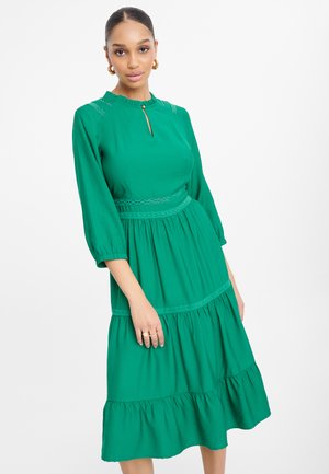Abito verde con un corpo aderente, scollatura rotonda e maniche a tre quarti. Presenta dettagli in pizzo testurizzato e strati nella gonna.