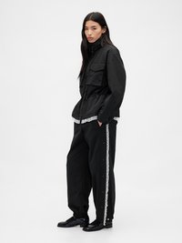 Sort jakke med lynlås og snoreliv, der har to brystlommer. Sorte bukser med snørebånd og trykknapper langs siderne.