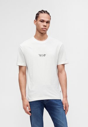 Jeune homme aux cheveux tressés portant un t-shirt blanc avec un petit motif noir et un jean bleu, debout devant un fond uni.
