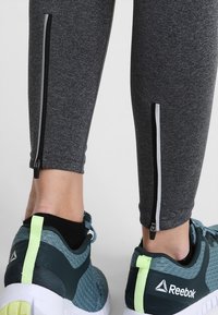 Gambe inferiori che indossano leggings grigio scuro con cerniere nere e sneakers Reebok blu con linguette al tallone verde neon e calzini neri alla caviglia.