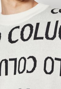 Suéter de punto blanco con gran texto negro en diferentes orientaciones. El texto dice "COLUC" y otras letras. Detalle de cuello acanalado visible.