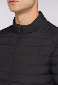 Černá prošívaná bunda s vysokým límcem a předním zipem. Obsahuje horizontální prošívání a decentní logo na hrudi. Hladká textura.