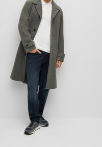 Šedý vlněný kabát s klopovým límcem, dvěma předními knoflíky a bočními kapsami, v kombinaci s tmavě modrými džínami a šedými teniskami.