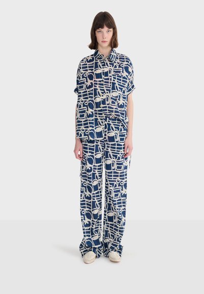Kobieta ubrana w luźną granatową koszulę i spodnie z białymi abstrakcyjnymi wzorami kratki i liści, stojąca na białym tle.