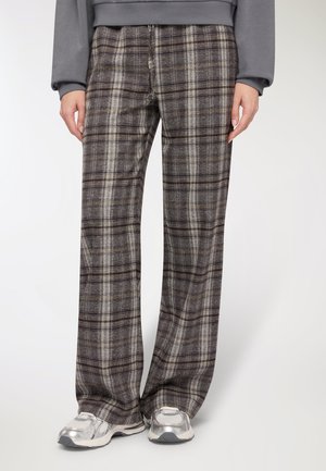 Pantaloni a quadri in tonalità grigio scuro e marrone con strisce crema, caratterizzati da una vestibilità ampia e una texture morbida, abbinati a sneakers argento.