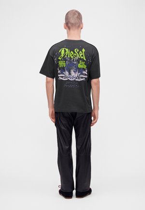 Mand med kort hår iført en sort grafisk Diesel T-shirt og sorte bukser, står med ryggen mod kameraet på en ensartet lys baggrund.
