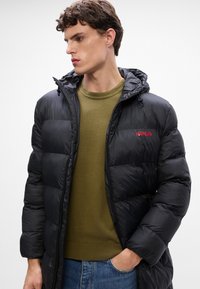Chaqueta tipo abrigo en negro con cierre delantero, diseño de acolchado y capucha con cordón. Hombros caídos y un logo rojo de "HUGO". Lleva puesta sobre un suéter verde.