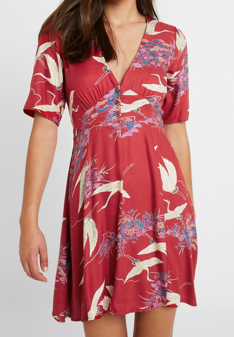 Femme portant une robe rouge à manches courtes avec des motifs blancs de grue et floraux et des boutons à l'avant sous un profond décolleté en V.