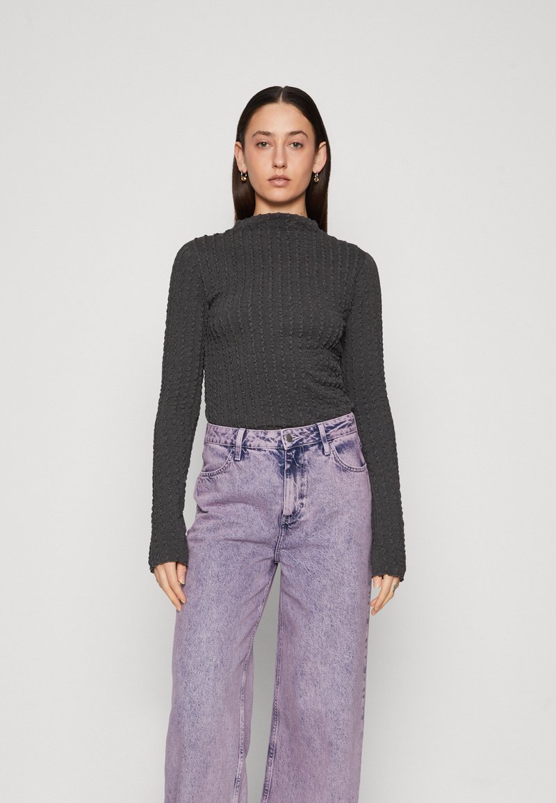 Mørk grå topp med lange ermer og strukturert overflate, høy hals, kombinert med høyt liv, vide lyse lilla jeans med en falmet vask.