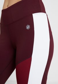 Viininpunaiset leggingsit valkoisella sivupaneelilla. Niissä on istuva malli, pehmeä koostumus, litteät saumat ja pieni logo ylemmässä reidessä.