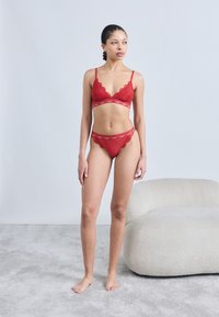 Rotes Spitzen-Bralette mit gewellten Kanten und passendem Spitzen-String, ausgestattet mit einem elastischen Band mit Markenlogo. Weicher, strukturierter Stoff.
