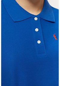 Niebieska koszulka polo z trzema białymi guzikami, ryflowanym kołnierzykiem oraz pomarańczowym haftowanym logo gracza polo po lewej stronie. Teksturowana tkanina.