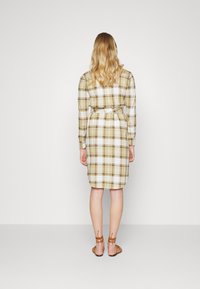 Abito camicia a maniche lunghe con un leggero motivo a quadri nei toni di verde, marrone e bianco. Include una cintura in vita e un orlo arrotondato; indossato con sandali marroni.